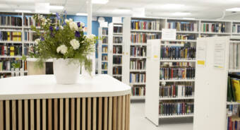 Kirjaston kirjahyllyjä joiden etualalla on kukkaruukku asiakaspalvelu tiskin kulmalla.