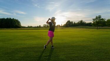 Auringonlaskussa golffaava nainen jolla vaaleanpunaiset shortsit ja musta paita.
