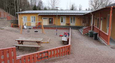 keltainen puinen päiväkodin rakennus jonka edessä on keinu, hiekkalaatikko ja pihapöytä.