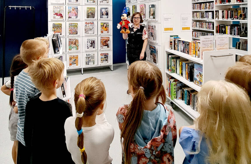 Esikoululaisia kirjastovierailulla, kirjastonhoitajalla Mikki Hiiri -pehmohahmo kädessä.
