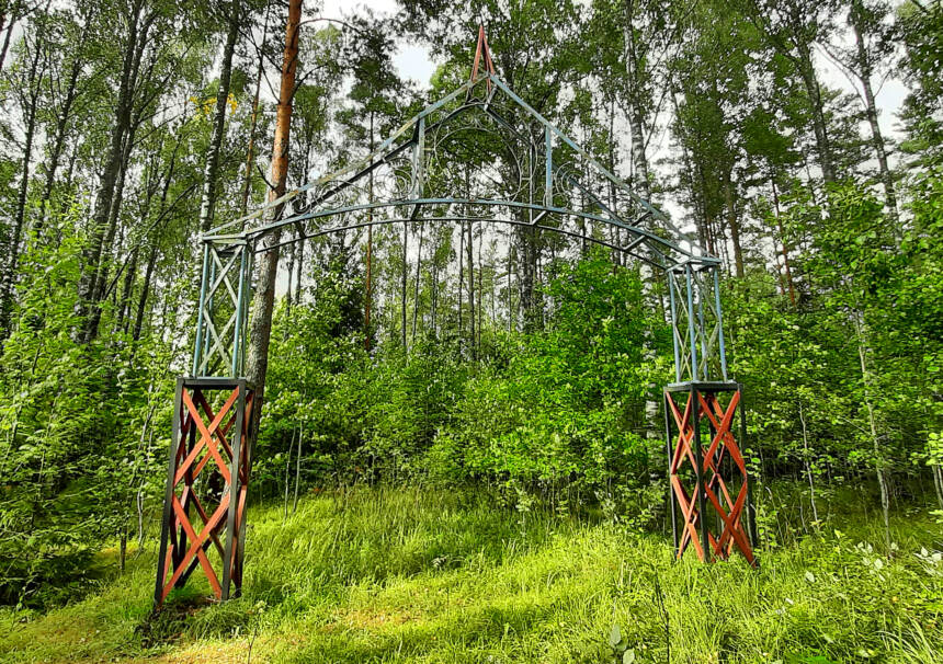 En triumfbåge i stål i en lövskog, röd till nedre delen och grå i den mellersta och övredelen.