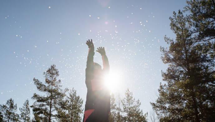 En person slänger upp snö mot solen och blå himmel.