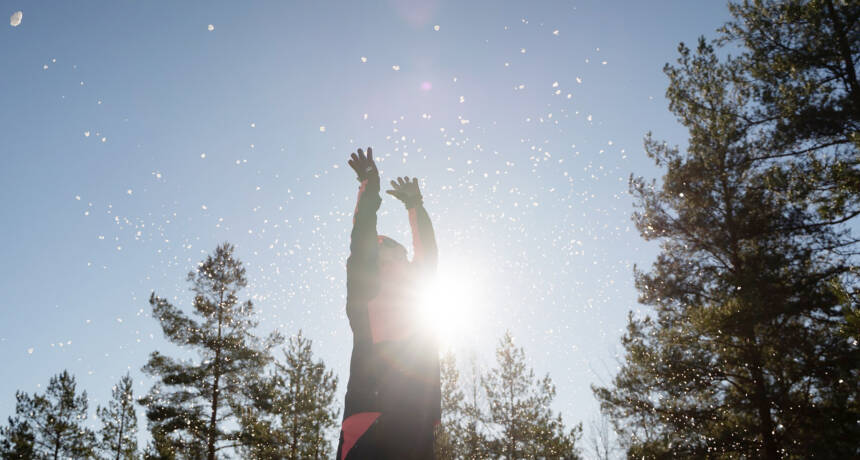 En person slänger upp snö mot solen och blå himmel.