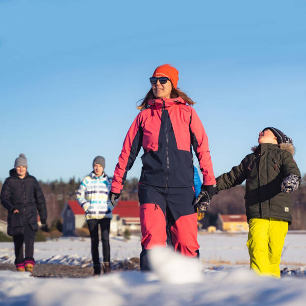 Äiti ja poika sekä muita ihmisiä taustalla kävelevät aurinkoisessa talvimaisemassa.