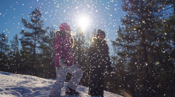 Två barn står på en snöig sluttning med solen i bakgrunden, i förgrunden yr snöflingor